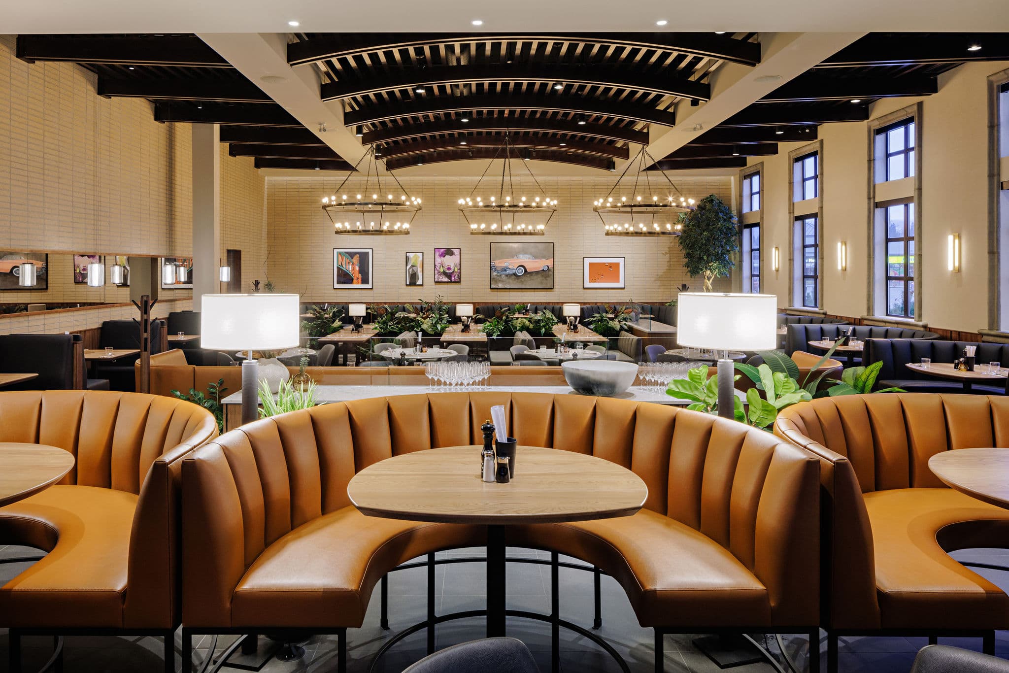 Restaurant interior with orange booths, wooden tables, plant accents, contemporary chandeliers, and natural light streaming through large windows.