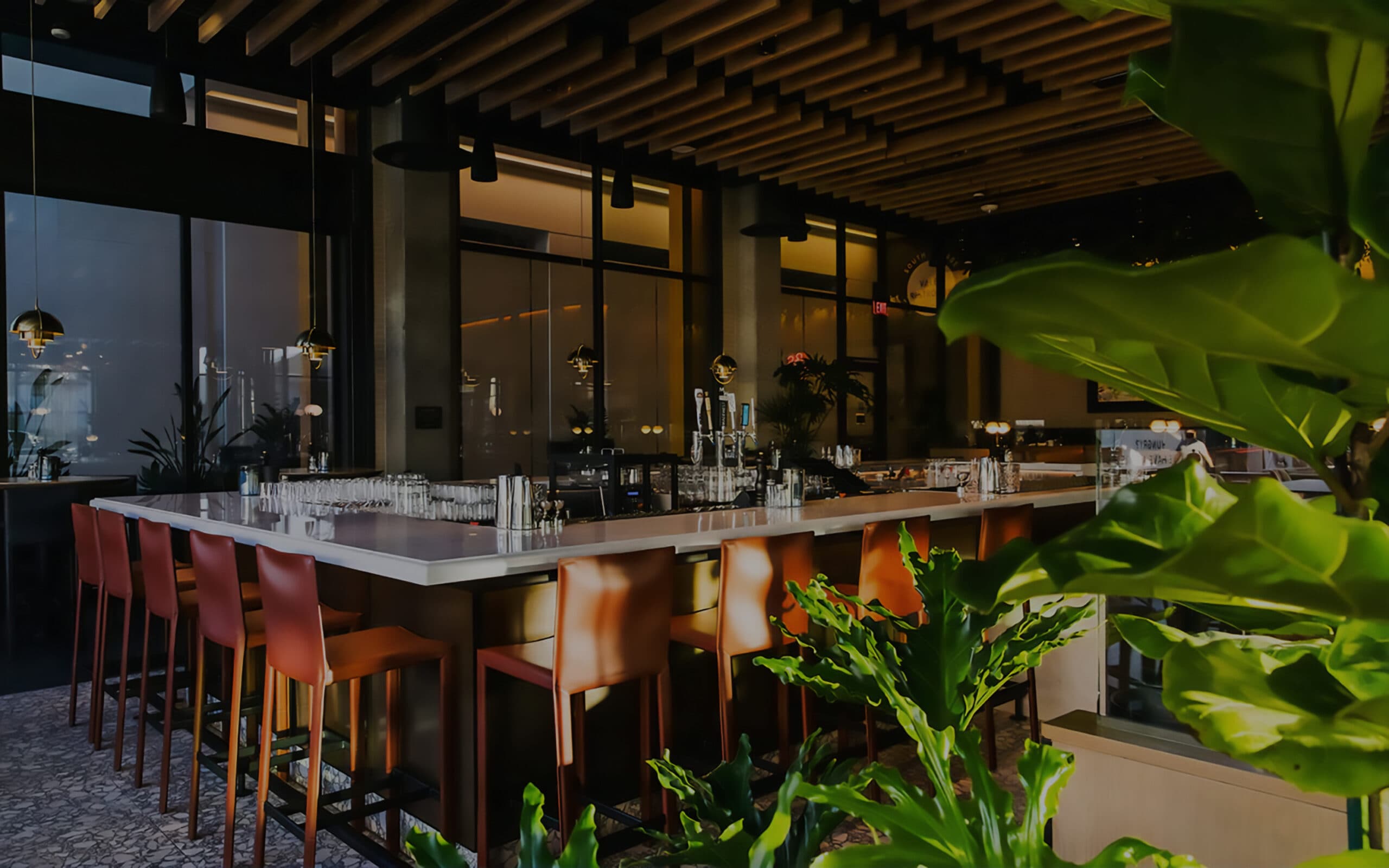 Restaurant interior of a bar with high stools, green plants, soft ambient lighting and natural light with large windows that overlook a lit hallway.