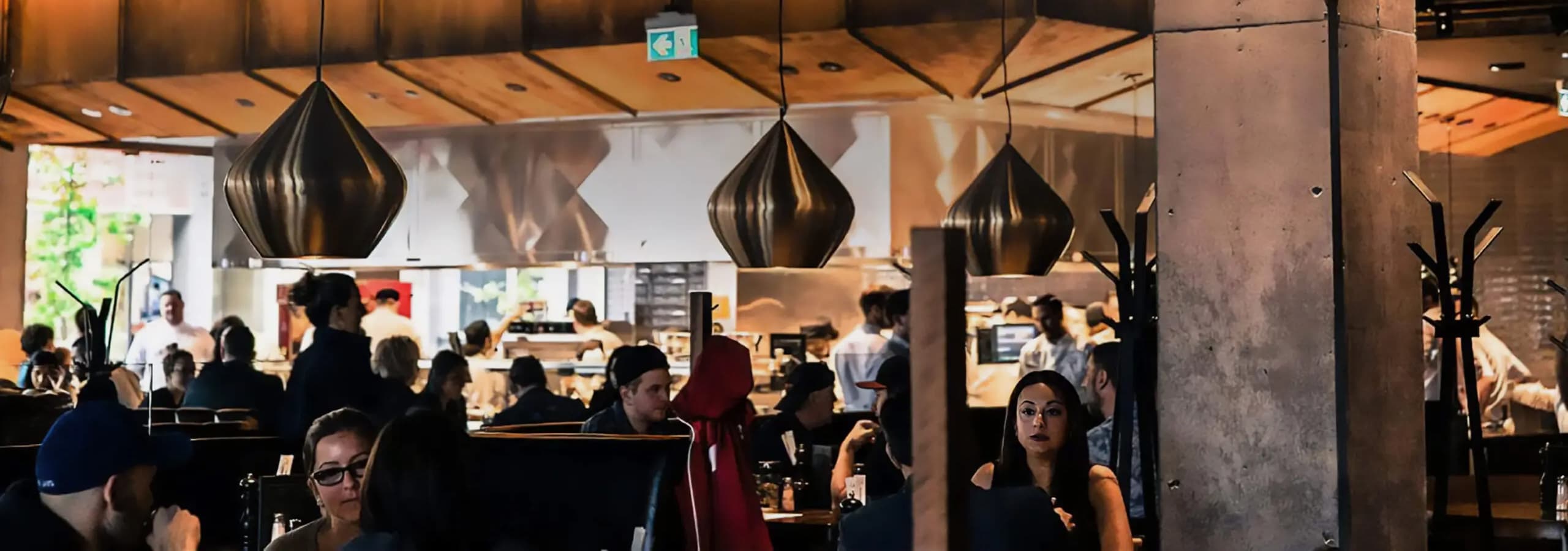 Busy restaurant interior with large light fixtures, people sat at tables and an open kitchen in the background.
